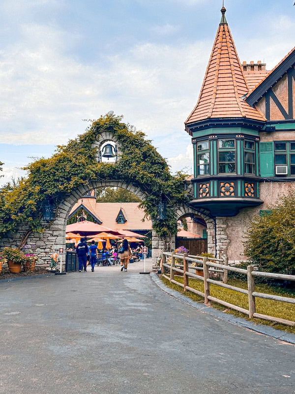 Entrance to the Bauernhof Courtyard at Grant’s Farm in St. Louis, featuring an ivy-covered stone archway and a turreted building with timber accents. Visitors walk through the arch toward the courtyard’s outdoor seating area with colorful umbrellas.