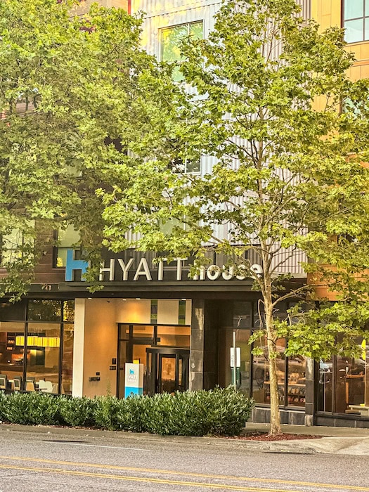 Front entrance of the Hyatt House Seattle with modern building exterior and leafy green trees framing the hotel.