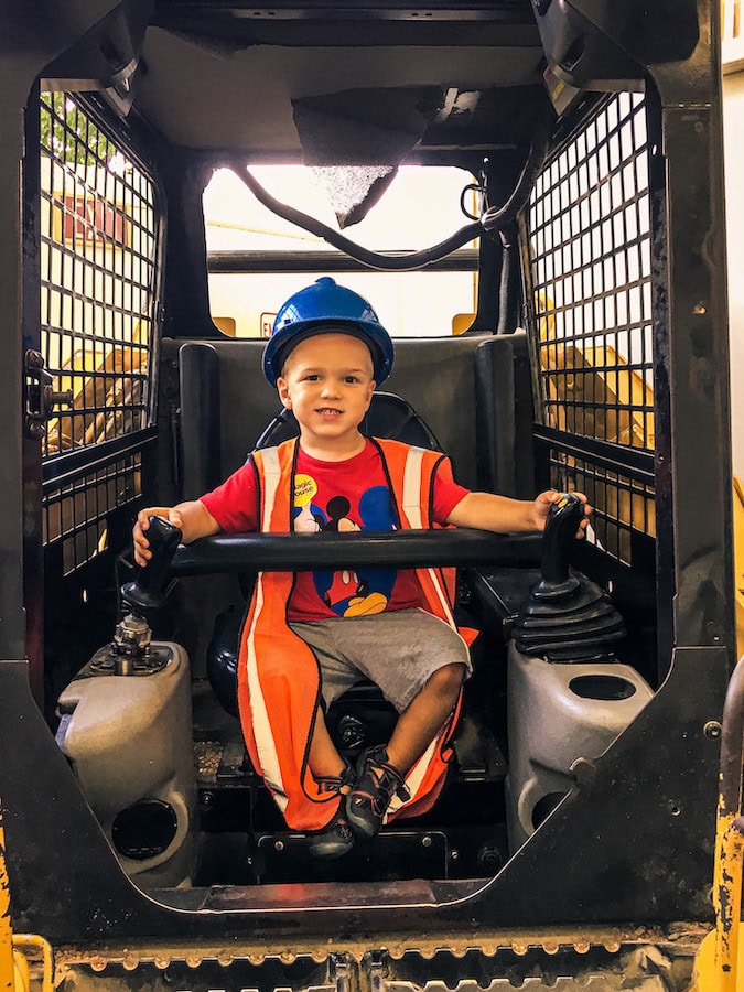 Child wearing a hard hat and safety vest sitting in a construction vehicle in the Construction Zone exhibit at The Magic House in St. Louis.