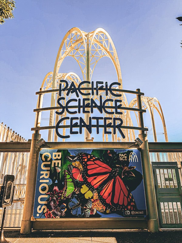 Entrance sign for the Pacific Science Center with arched white structures and a colorful “Be Curious” butterfly mural below.