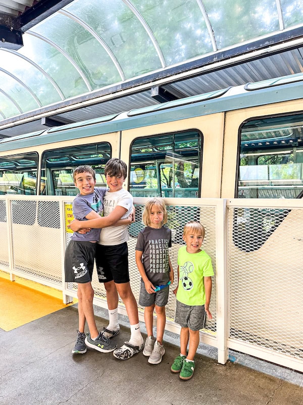 Four children standing and smiling on the platform next to the Seattle Monorail, with the train visible behind them under a curved glass canopy.