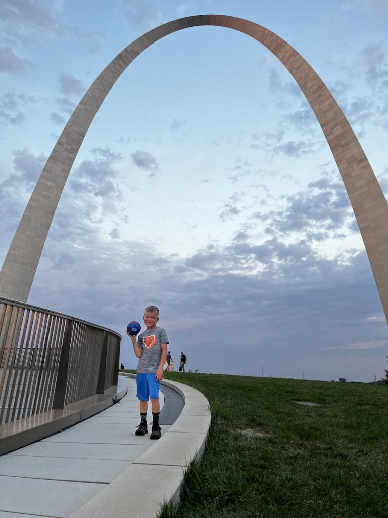 A young boy holding a small blue soccer ball stands on a curved walkway near the Gateway Arch in St. Louis at sunset, with the Arch towering above him against a pastel sky.