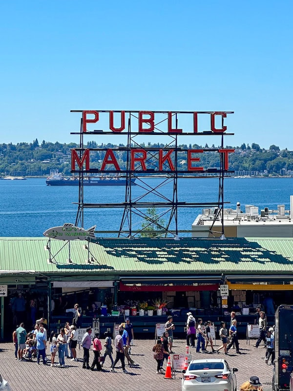 The iconic red “Public Market” sign at Pike Place Market in Seattle, with the waterfront and distant hills in the background and crowds walking below.