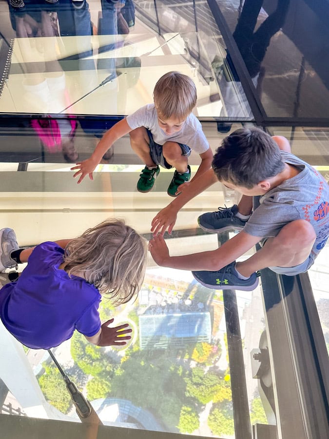 Three children sitting and crawling on the rotating glass floor observation deck of the Space Needle, looking down at the view below