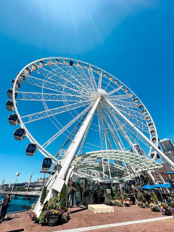 A low-angle view of the Seattle Great Wheel on the waterfront, with bright blue sky and sunlight shining behind the Ferris wheel.