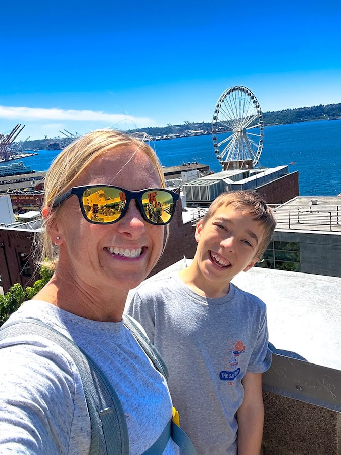 An adult and a child smiling for a selfie with the Seattle waterfront, Seattle Great Wheel and blue water in the background.