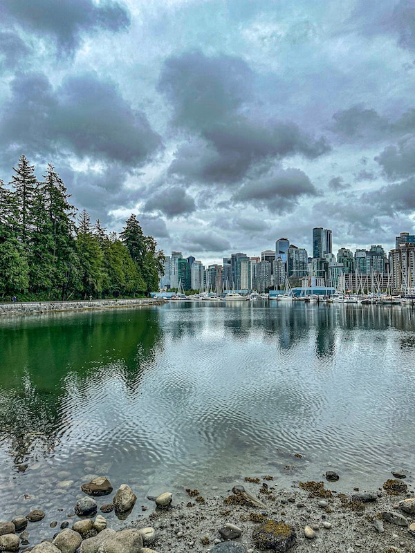 View of downtown Vancouver’s skyline from the Stanley Park seawall, with sailboats in the marina and calm water reflecting the cloudy sky and surrounding trees.