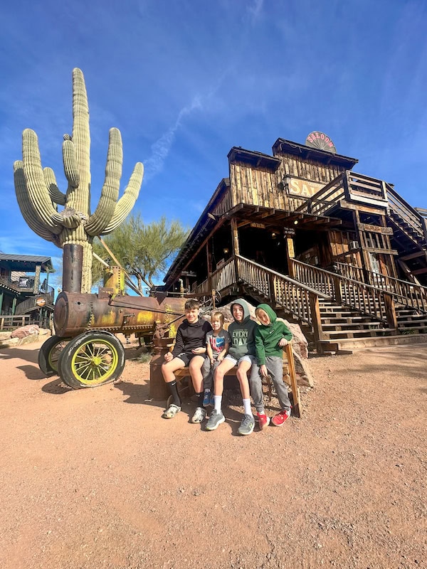 Kids sitting on a bench near a cactus and vintage train display with a rustic looking saloon in the background.