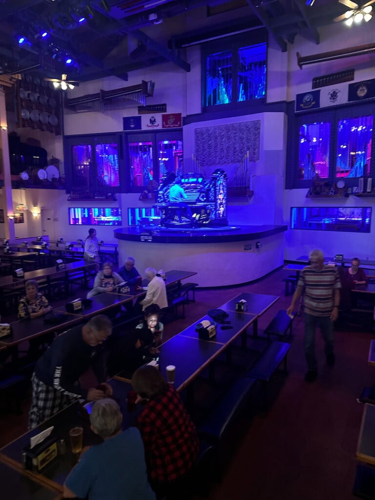 Dining room at Organ Stop Pizza with guests seated at long banquet-style tables while the organist performs on the central stage.