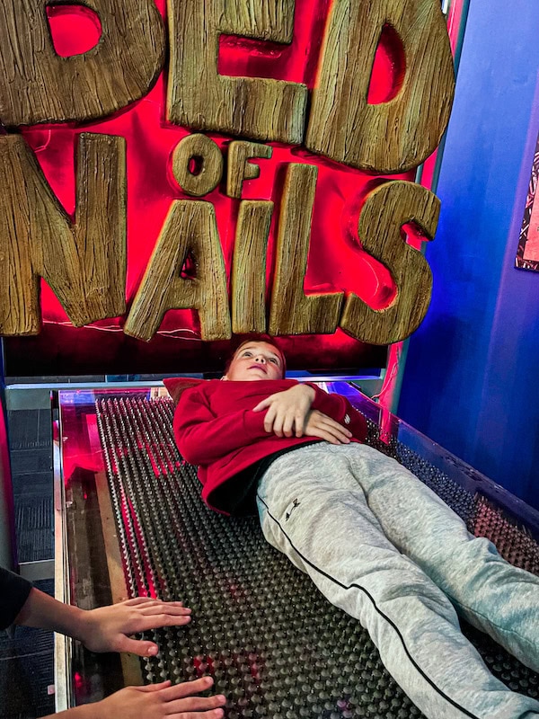 Child lying on the &ldquo;bed of nails&rdquo; exhibit inside WonderWorks in Branson, Missouri, with bright exhibit signage in the background.