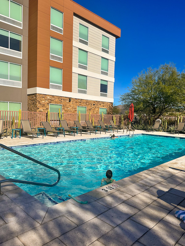 Outdoor pool at Home2 Suites Mesa Longbow in Arizona with lounge chairs, desert landscaping, and kids swimming on a sunny day.