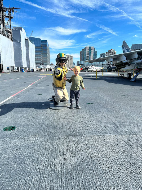 Little boy standing next to a pilot statue on the flight deck of the USS Midway Museum with downtown San Diego skyline in the background.