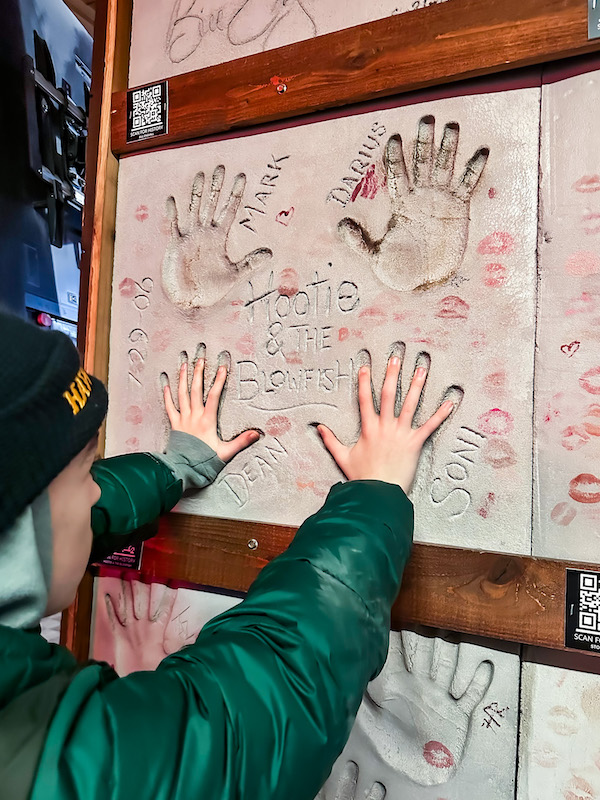 Boy wearing a green jacket and stocking hat placing his hands on the handprint of a famous performer handprints inside Billy Bob&rsquo;s Texas