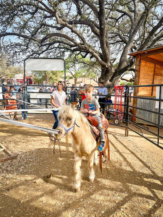 Child riding a small pony in a fenced dirt arena at the Fort Worth Stockyards with parents and other families nearby