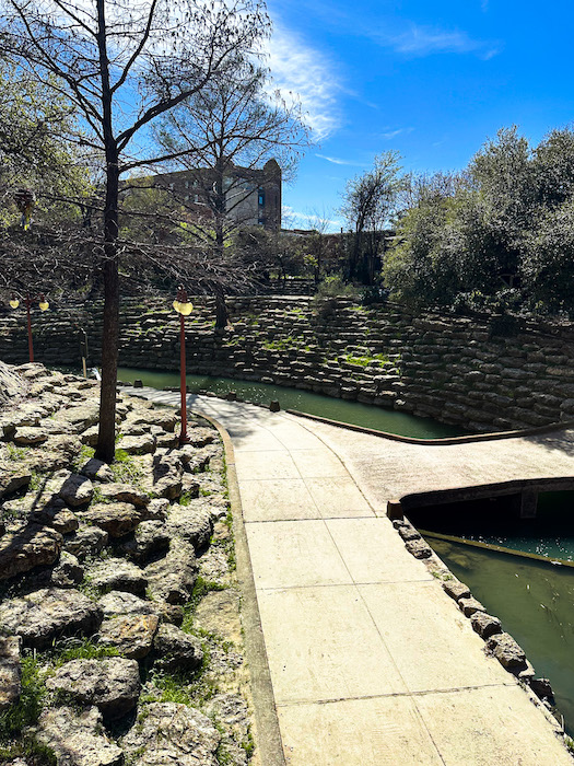 Paved walking path along Marine Creek with stone walls, trees, and water creating a quiet scenic area near the Fort Worth Stockyards