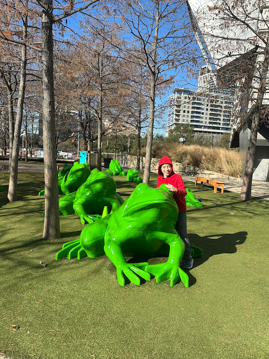 Child posing on bright green frog sculptures outside the Perot Museum of Nature and Science in Dallas, Texas, with city buildings in the background