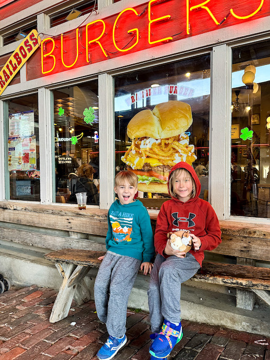 Two kids sitting on a bench outside Trailboss Burgers at the Fort Worth Stockyards with a giant burger display visible through the window