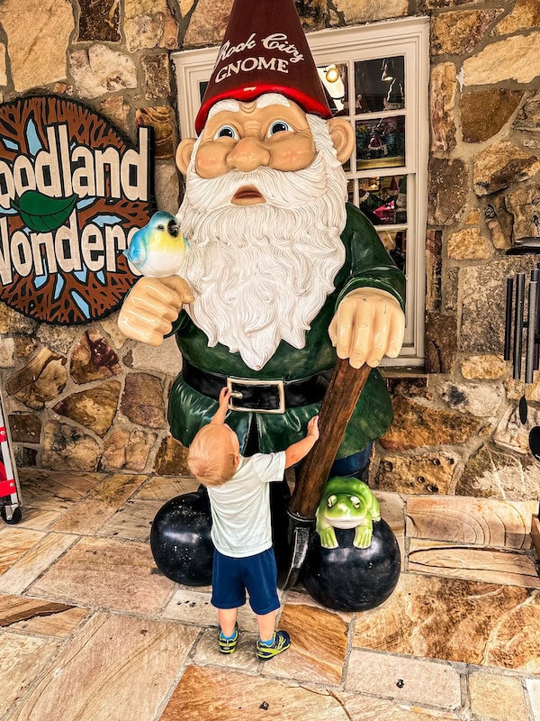 Child exploring a large Rock City gnome statue with a red hat, bird, and frog outside of a stone building
