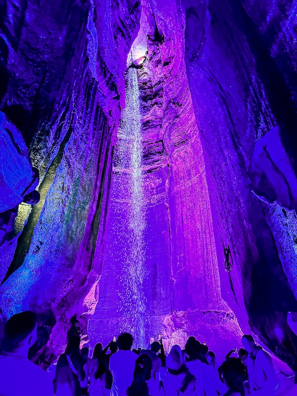 People looking at a waterfall in a cave with pink and blue colored lights highlighting the area