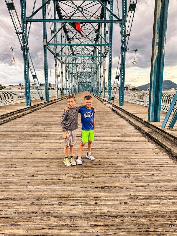 Two boys with their arms around each other standing in the middle of a pedestrian bridge.