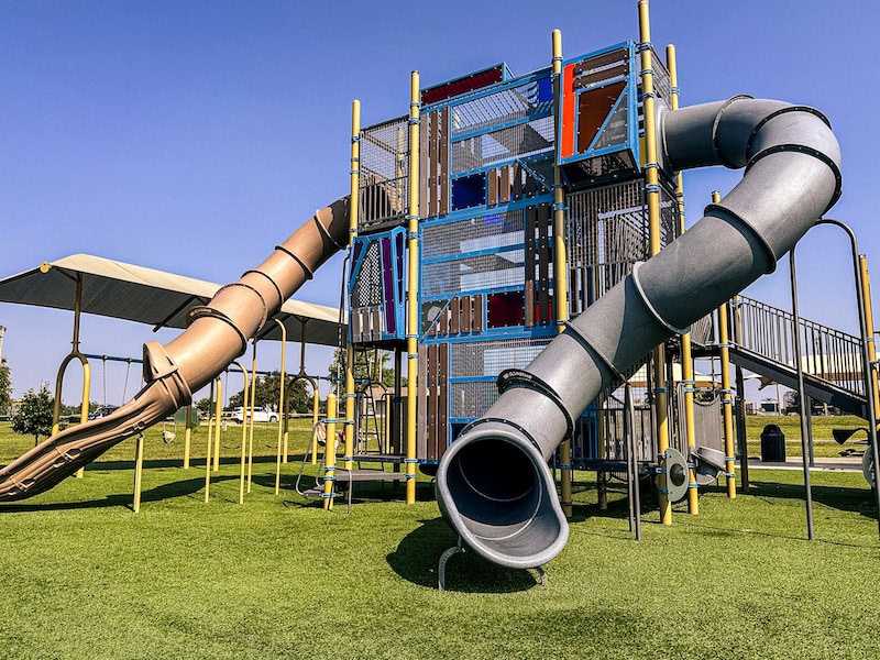 Large climbing playground with colorful towers, enclosed tube slides, and artificial turf at Centennial Park in Waukee, Iowa.