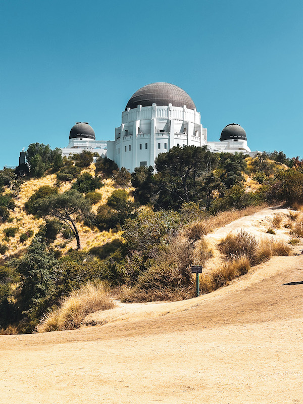 Griffith Observatory viewed from a hiking trail surrounded by dry hillsides and vegetation in Griffith Park.