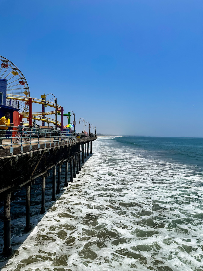 View from the Santa Monica Pier overlooking the Pacific Ocean, waves crashing below, and amusement rides visible along the pier.