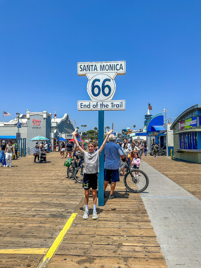 Child posing beneath the Route 66 End of the Trail sign on the Santa Monica Pier in California.