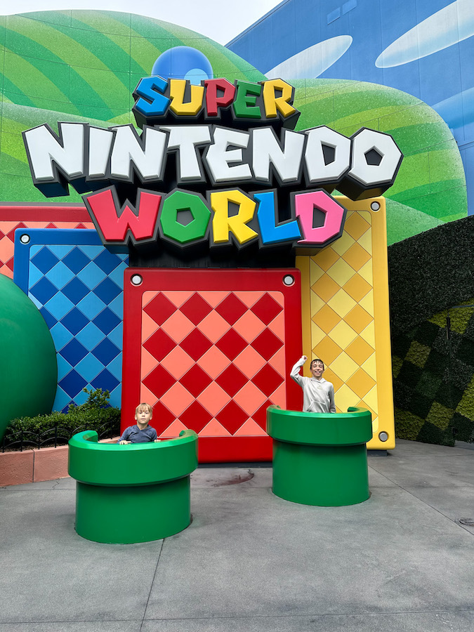Two boys posing in green warp pipes in front of the colorful Super Nintendo World entrance sign at Universal Studios Hollywood.