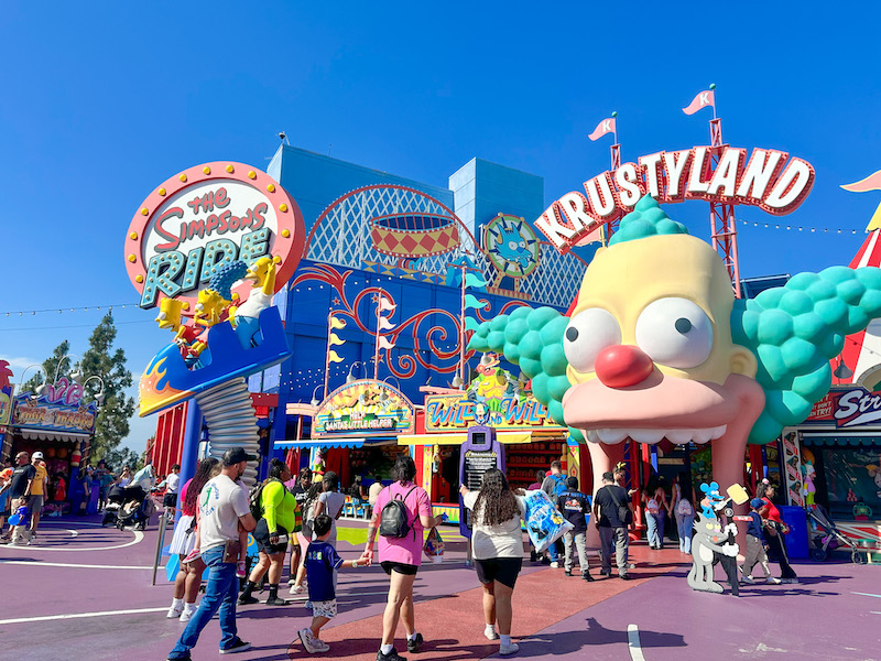 Krustyland entrance and The Simpsons Ride area at Universal Studios Hollywood with colorful carnival-themed buildings, Krusty the Clown's giant face, and park guests walking through the attraction.
