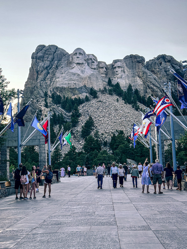 Mount Rushmore National Memorial viewed from the Avenue of Flags at dusk with visitors walking toward the presidential sculpture in South Dakota.