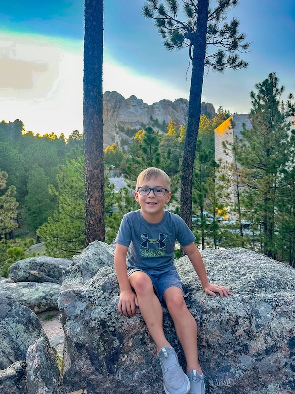 Child sitting on a rock with Mount Rushmore visible through the trees in the background in South Dakota.