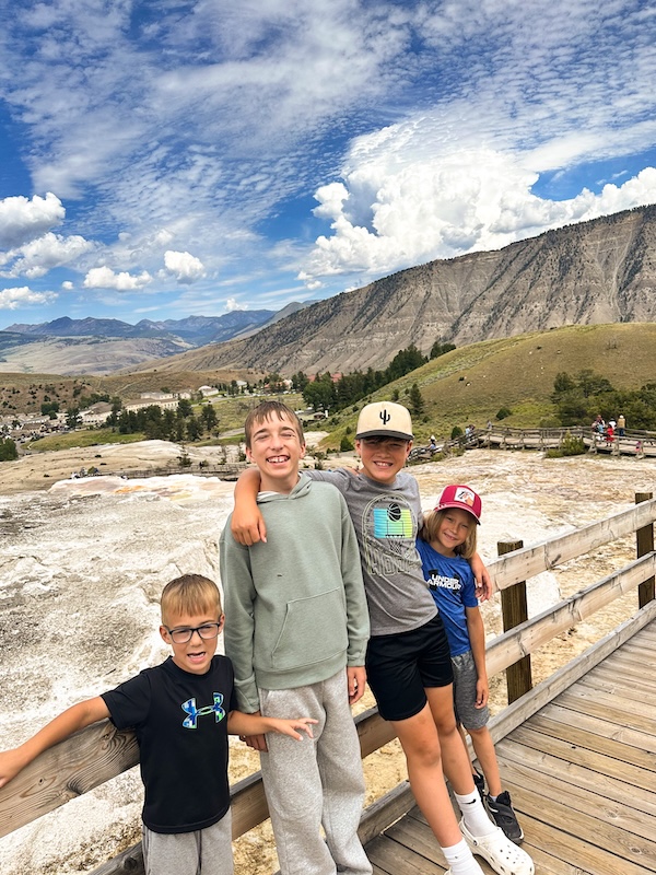 Four boys standing together on the boardwalk at Mammoth Hot Springs with the terraces and mountain scenery behind them in Yellowstone National Park.