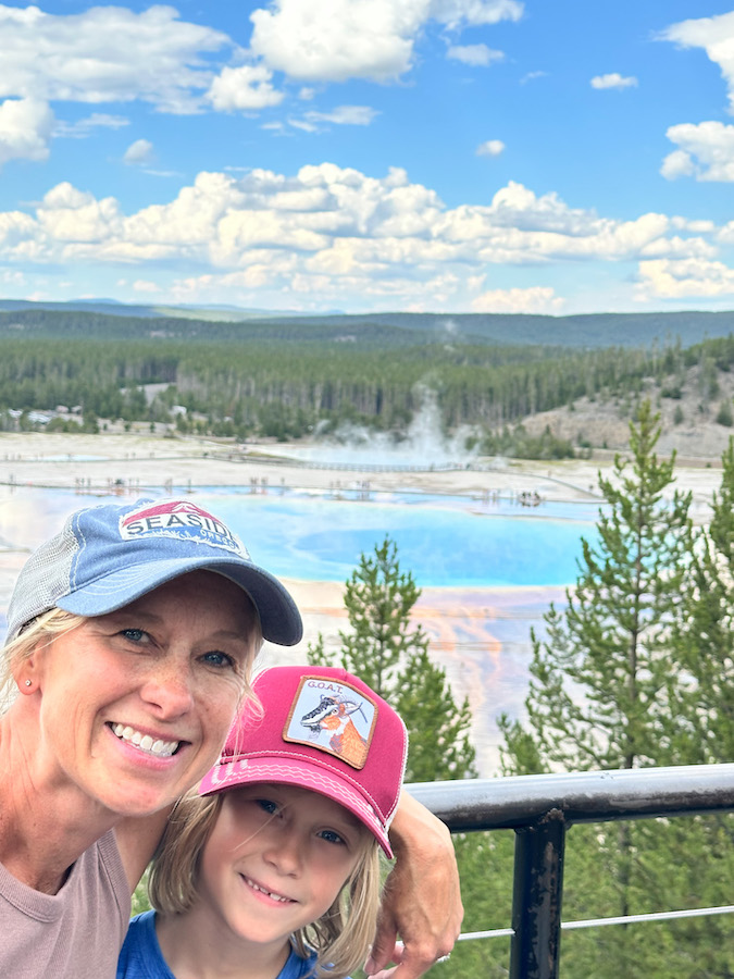 Mom and child smiling at the Grand Prismatic Spring Overlook with the colorful blue and orange spring behind them in Yellowstone National Park.