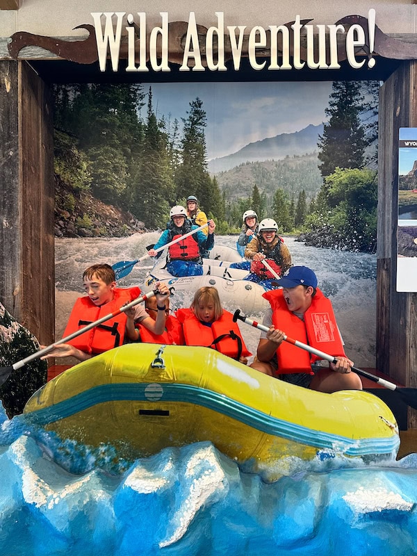 Three boys wearing life jackets and holding paddles in an interactive whitewater rafting display at the Northeast Wyoming Welcome Center.