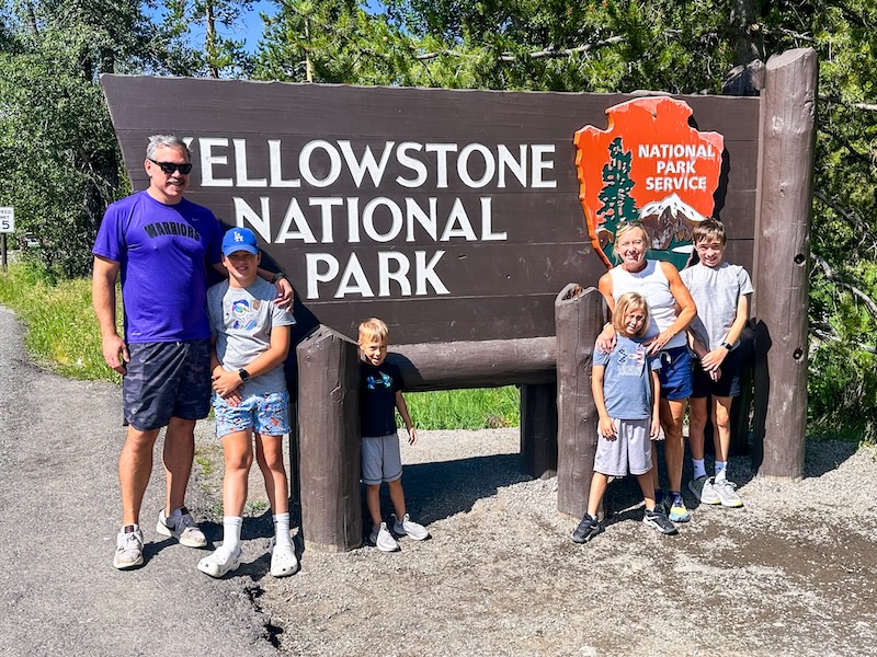 Family of six posing beside the Yellowstone National Park entrance sign on a sunny summer day.